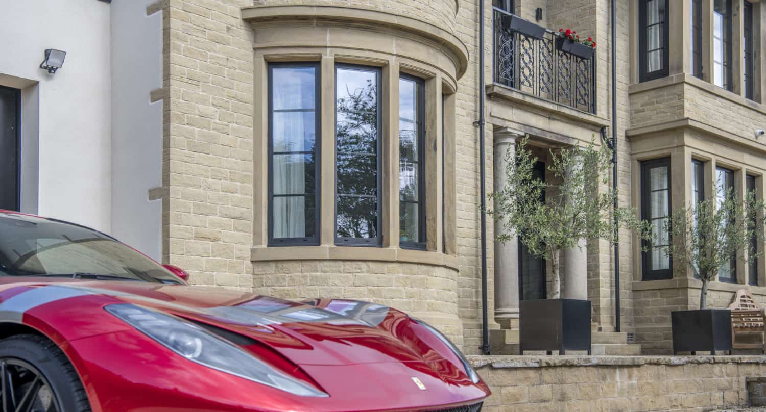 A shiny red Ferrari is parked in front of a large, elegant stone house with bay windows and a balcony adorned with flowers, featuring modern bifold doors that open onto the garden.