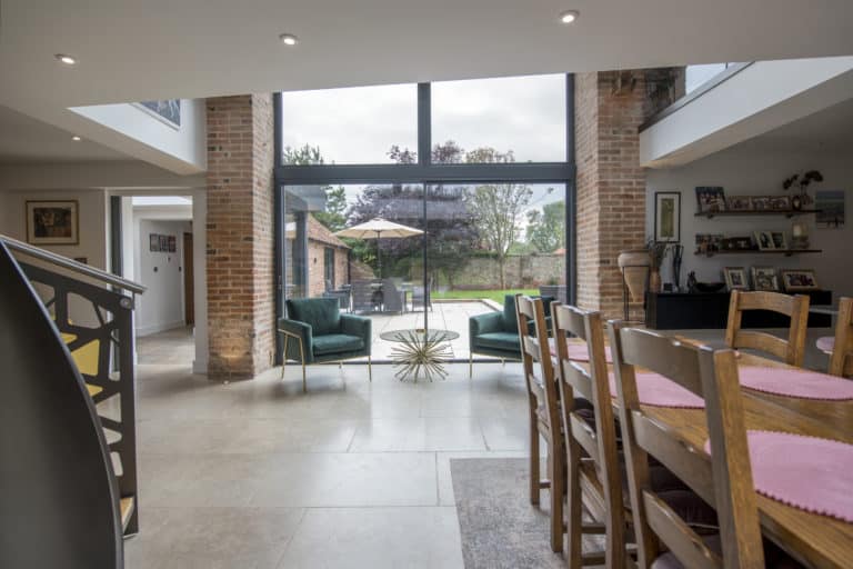 Modern dining and living area with large bespoke glass sliding doors opening to a patio. Wooden dining table with chairs, green armchairs by a gold coffee table, brick walls, and outdoor seating visible through the windows.