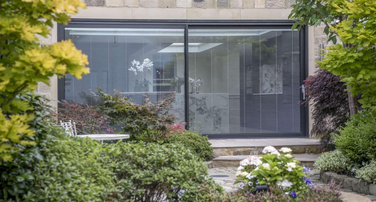 View of a modern glass sliding door with bespoke glass solutions on a stone building, surrounded by a lush garden with green bushes, colorful flowers, and a small stone pathway in the foreground.