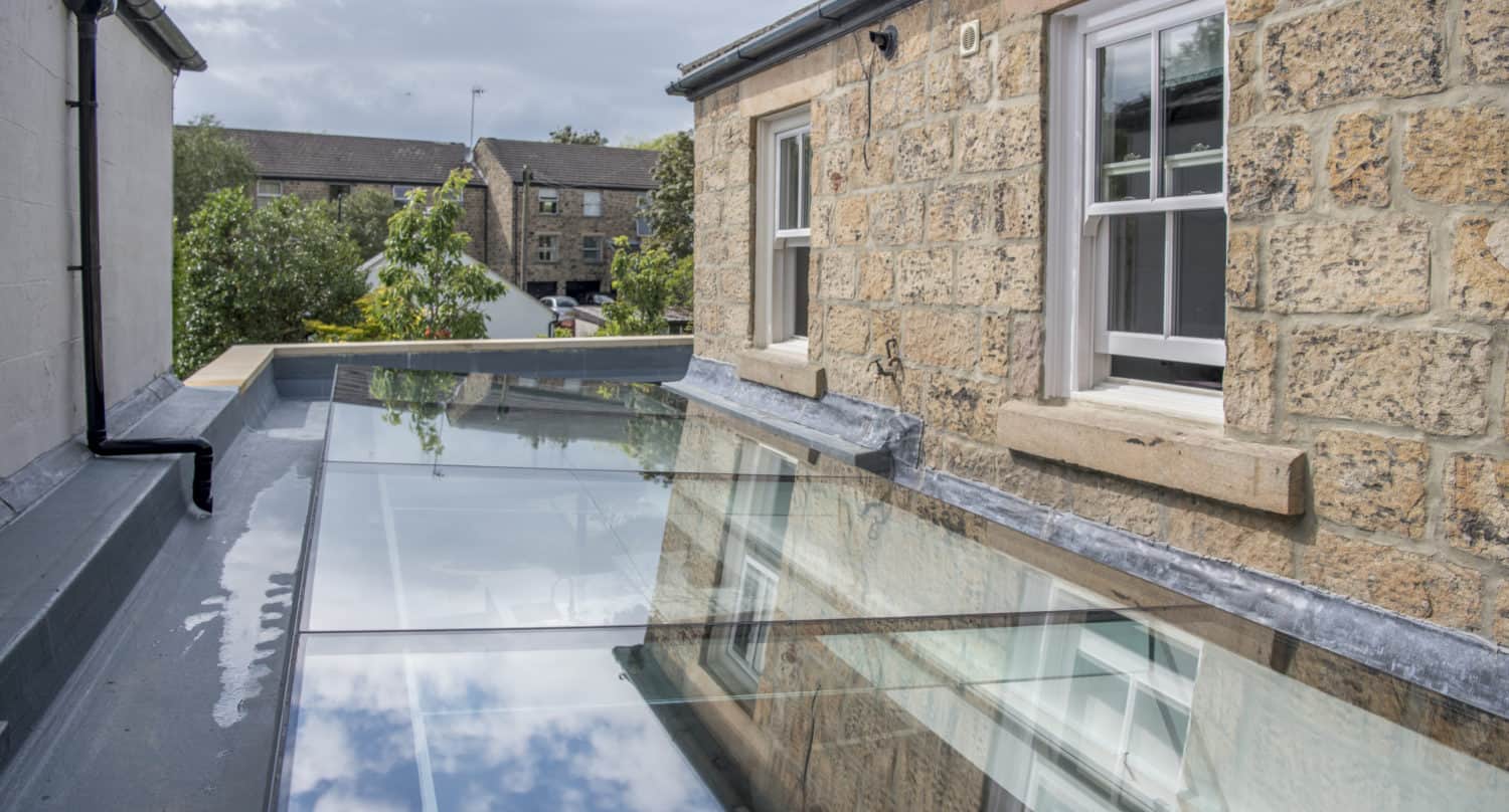 A modern glass roof extends between two stone buildings, reflecting the sky and clouds above. Sliding doors open onto the glass structure, while windows overlook lush greenery in the background.