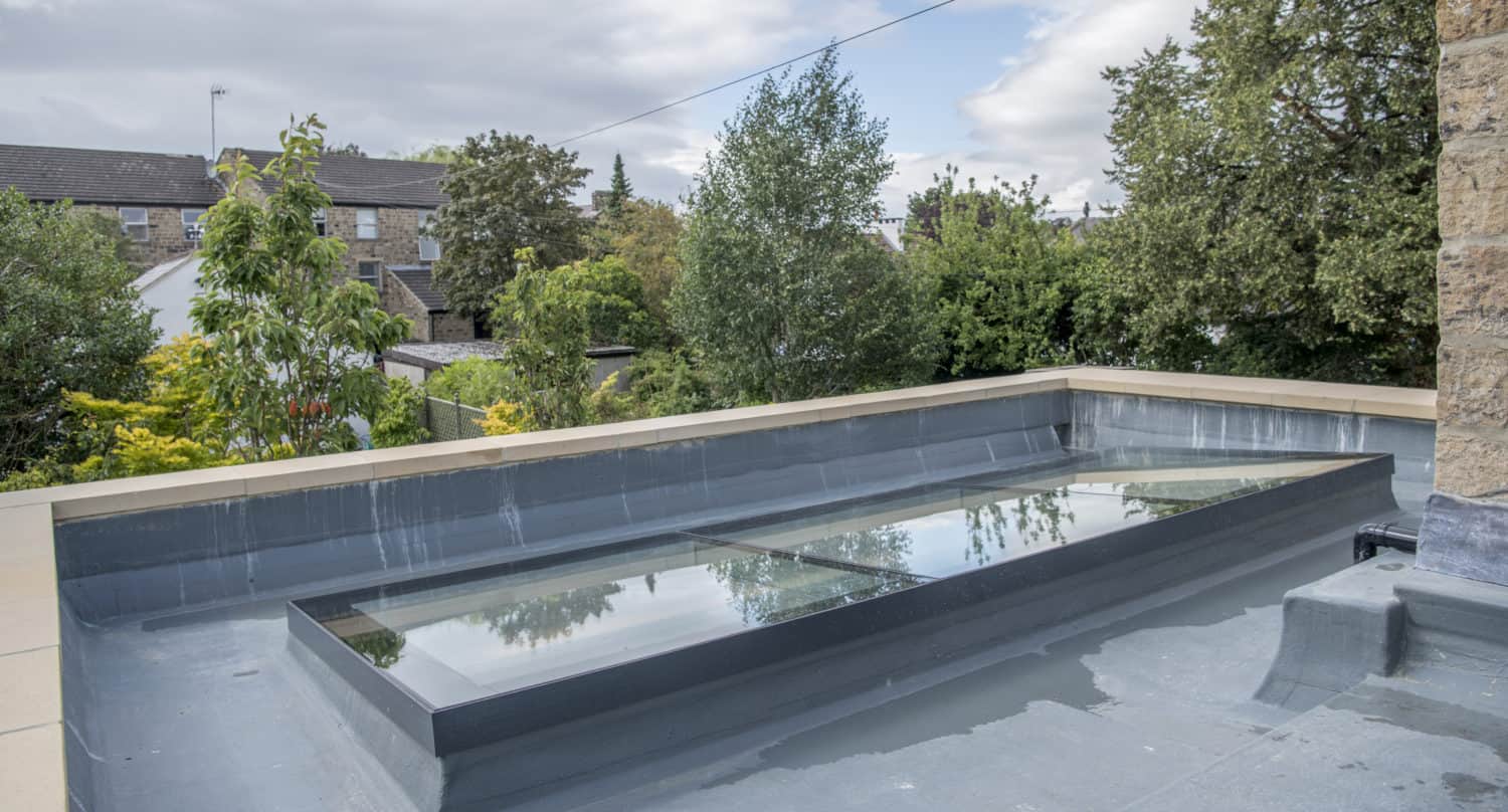 A flat rooftop with a rectangular glass skylight, showcasing bespoke glass solutions, surrounded by trees and neighboring houses under a partly cloudy sky.