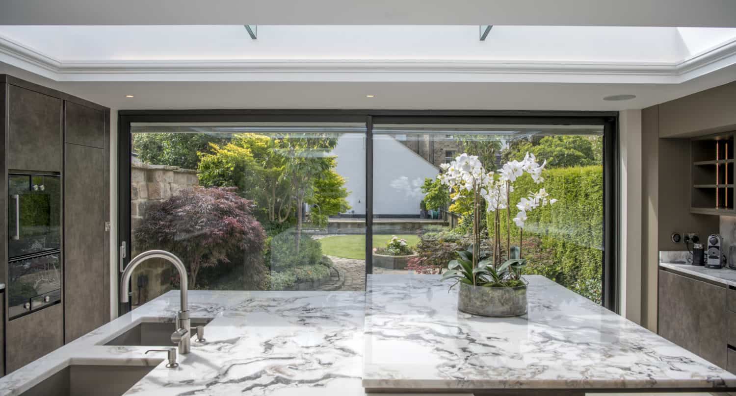 Modern kitchen with a marble island countertop, built-in sink, and potted white orchids. Large sliding doors open to a lush, green garden, letting in natural light from a skylight above.