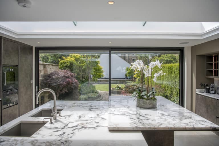 Modern kitchen with a marble island countertop, built-in sink, and potted white orchids. Large sliding doors open to a lush, green garden, letting in natural light from a skylight above.