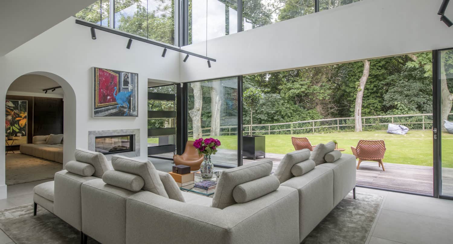 Modern living room with large floor-to-ceiling bifold doors, a light gray sectional sofa, a glass coffee table with flowers, wall art, and views of a green garden with outdoor seating.