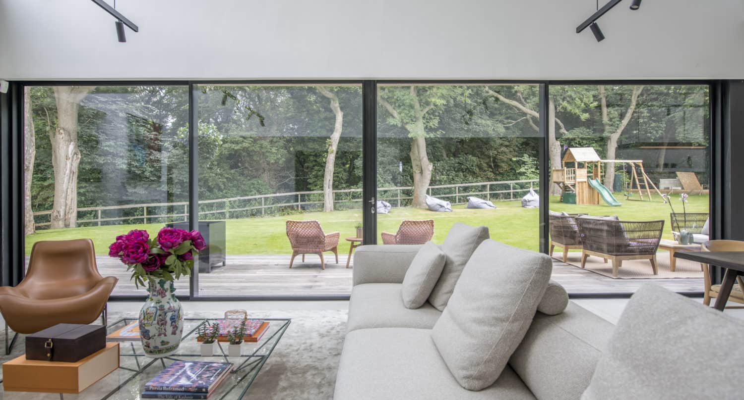 Modern living room with large glass windows and bespoke glass solutions overlooking a green backyard with trees, a wooden deck, outdoor chairs, a playset, and vibrant flowers on the coffee table in the foreground.