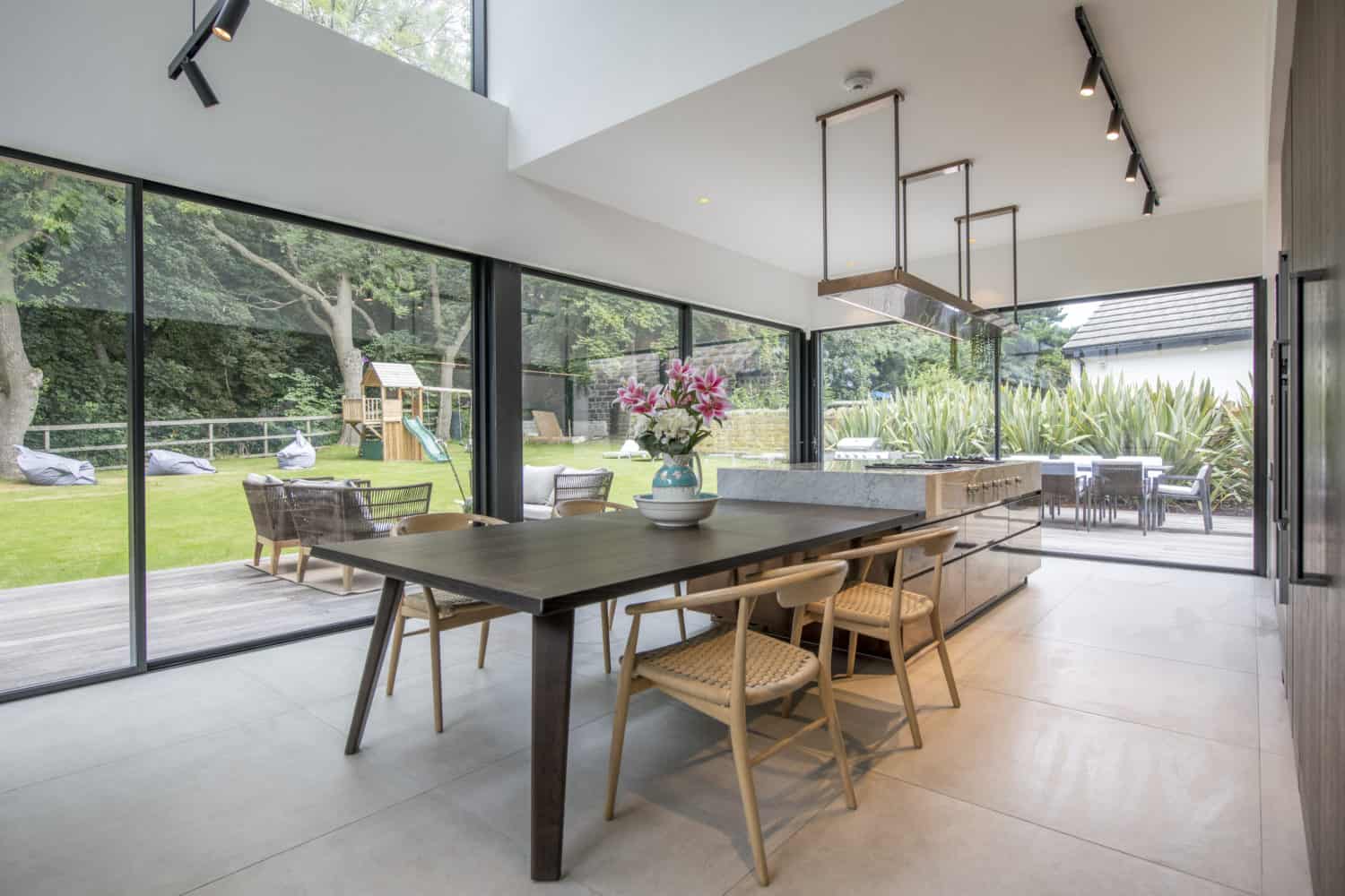 Modern dining area with large glass walls and bifold doors, a wooden table and chairs, a vase of pink flowers, and views of a green garden with a playset. Bespoke glass solutions fill the space with natural light.