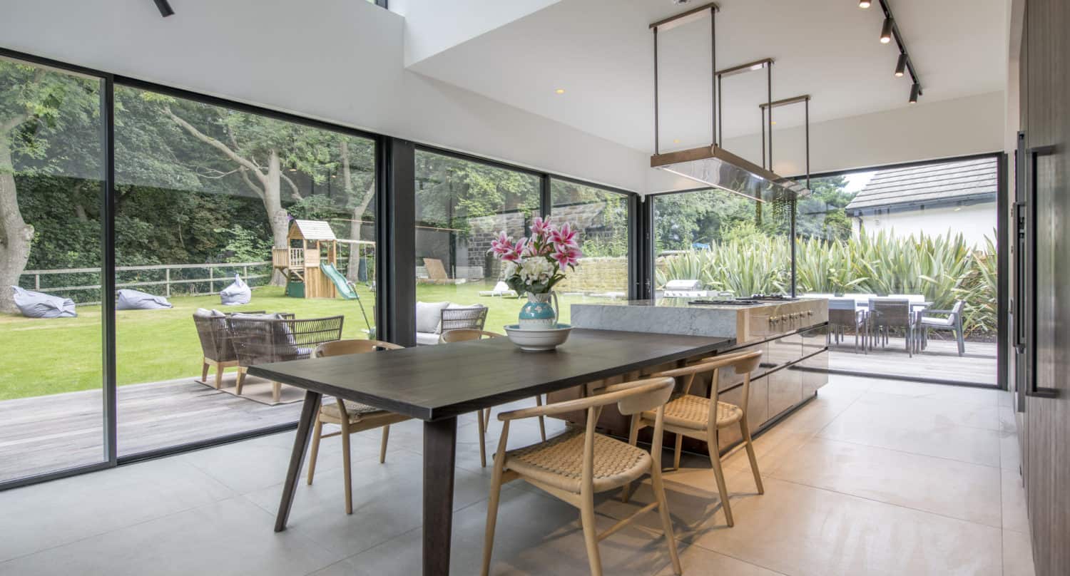 Modern dining area with large glass walls and bifold doors, a wooden table and chairs, a vase of pink flowers, and views of a green garden with a playset. Bespoke glass solutions fill the space with natural light.