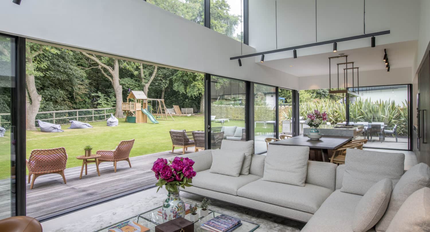 Modern living room with large glass walls and bifold doors overlooking a lush garden, patio seating, and a children’s playset. The interior features a white sectional sofa, glass coffee table, and elegant dining area.