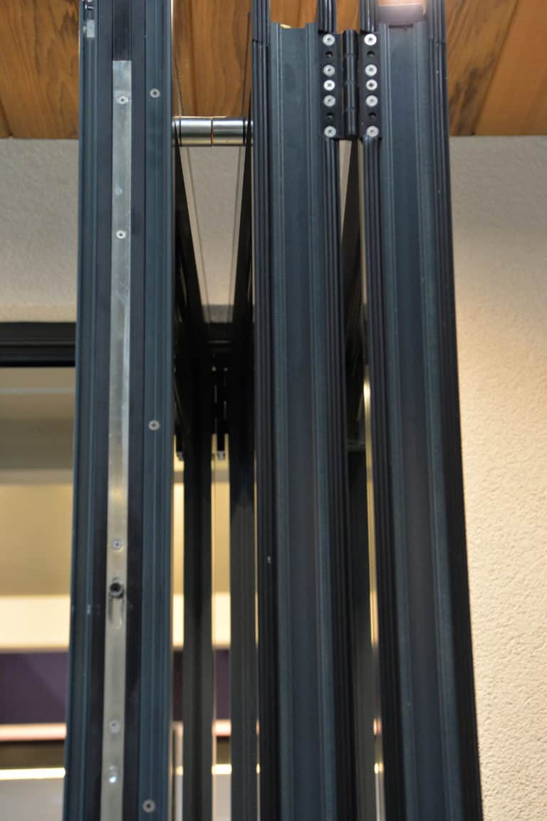 Close-up view of the hinge and track mechanism of a modern black aluminum folding door, mounted on a wooden ceiling and a light-colored wall, showcasing how bespoke glass solutions can seamlessly integrate with contemporary design.