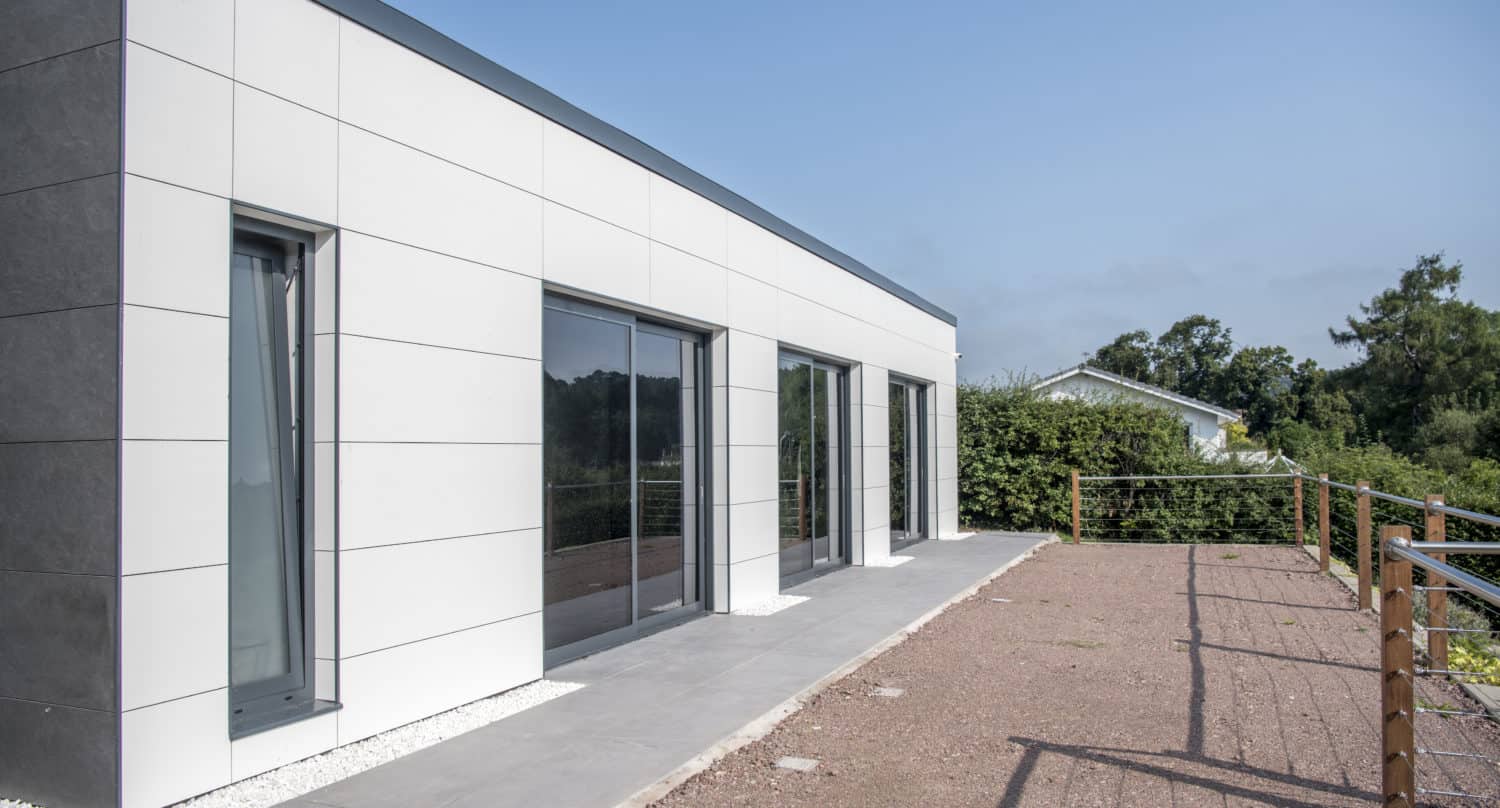 Modern single-story house with large bifold doors and a white and gray minimalist exterior, set on a paved patio with a gravel area and surrounded by greenery under a clear blue sky.