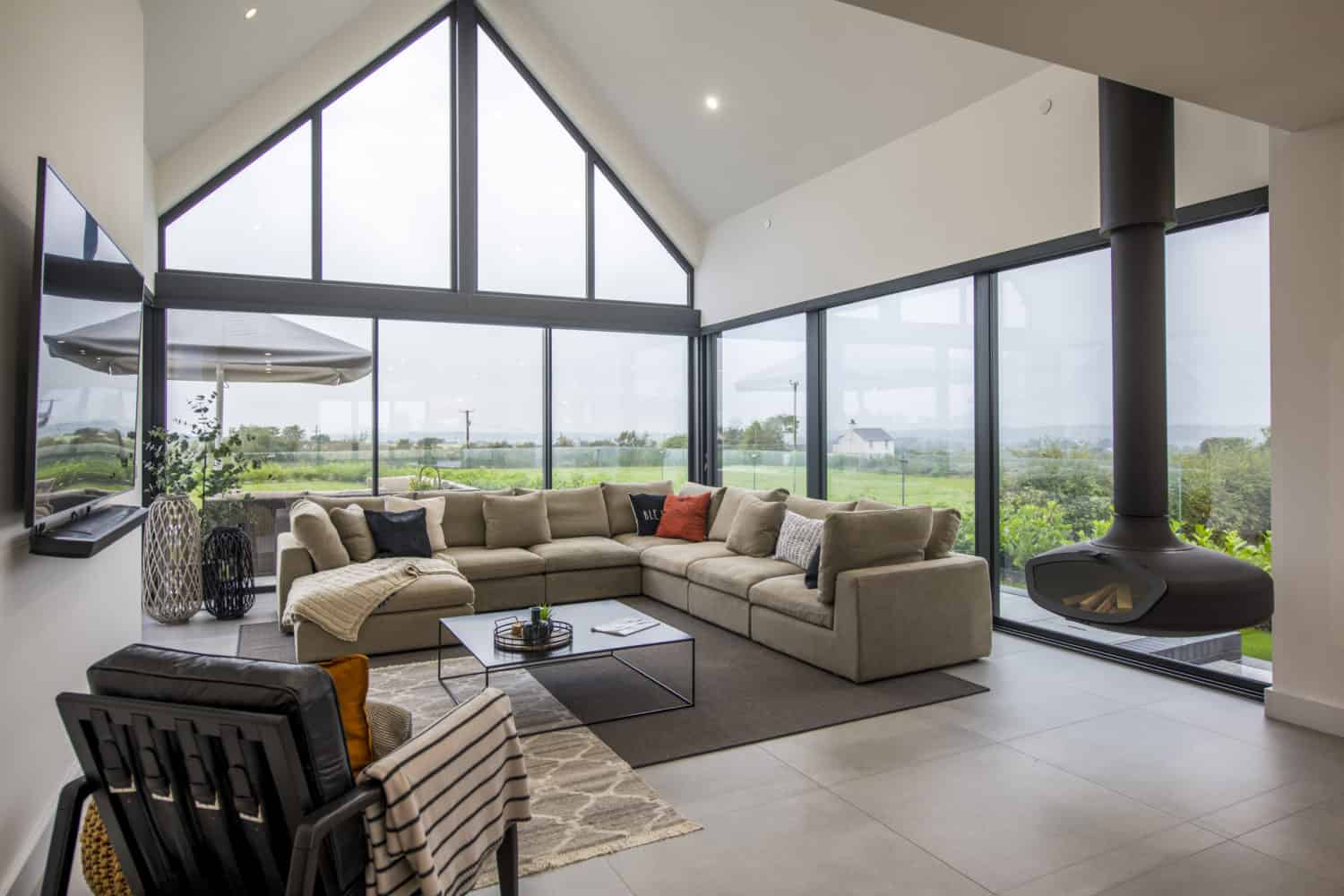 A modern living room with large floor-to-ceiling bespoke glass solutions, a beige sectional sofa, a black suspended fireplace, and views of green fields outside. The room features minimalist decor and abundant natural light.