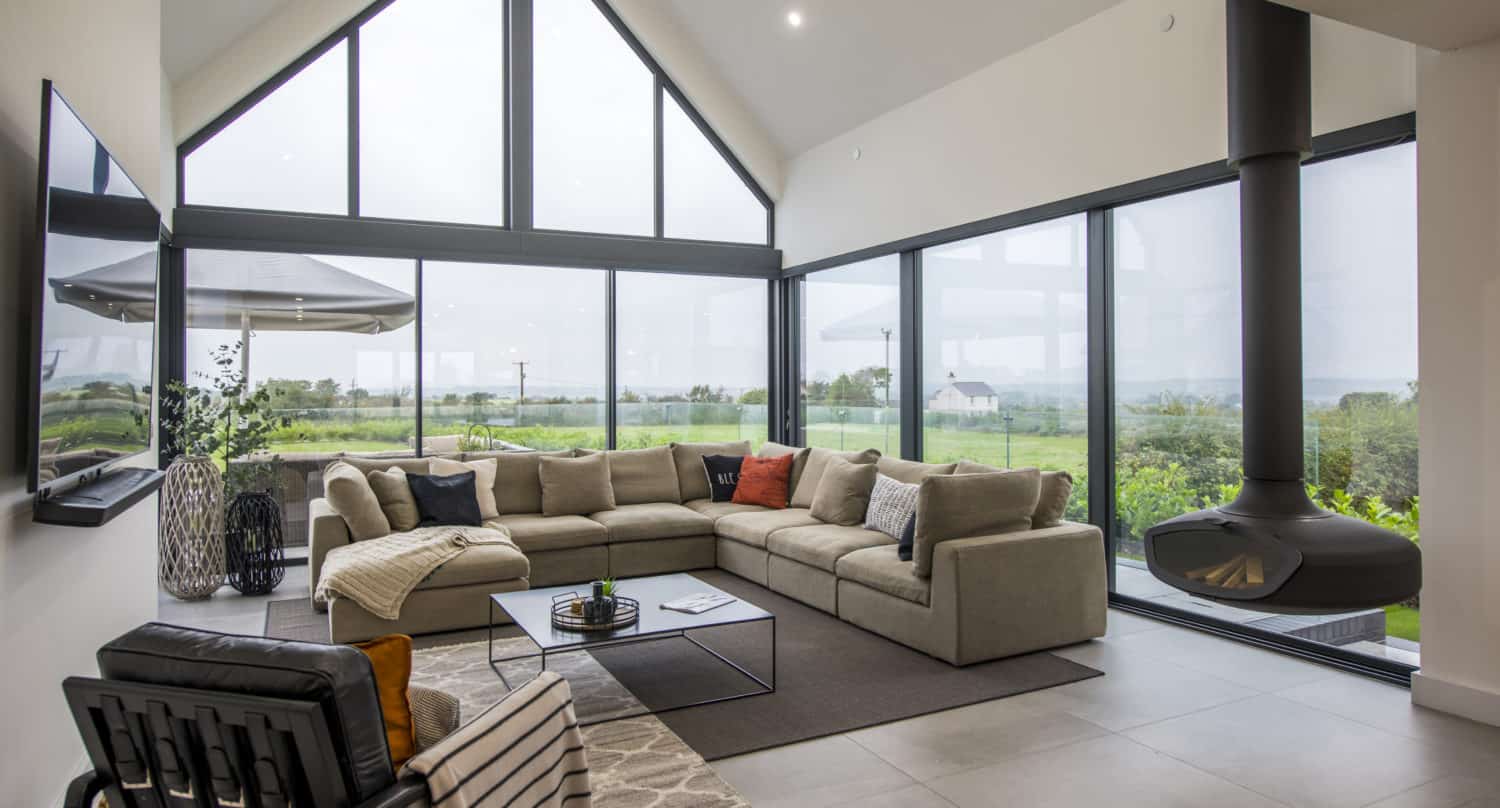 A modern living room with large floor-to-ceiling bespoke glass solutions, a beige sectional sofa, a black suspended fireplace, and views of green fields outside. The room features minimalist decor and abundant natural light.