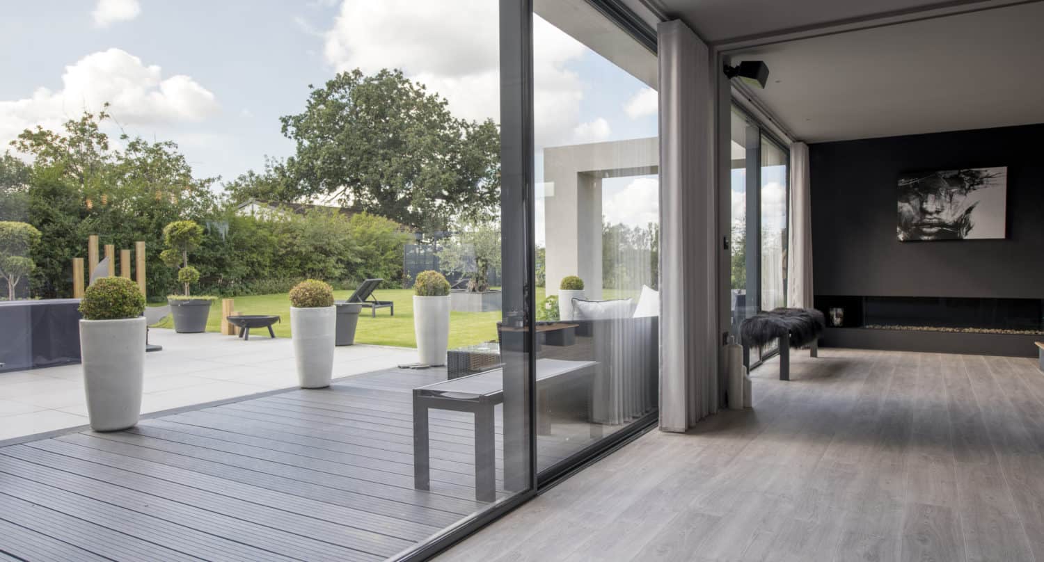 Modern living room with large bifold doors opening to a patio and garden. The room has light wood flooring and minimal furnishings; outside, large potted plants and a green lawn are visible under a partly cloudy sky.
