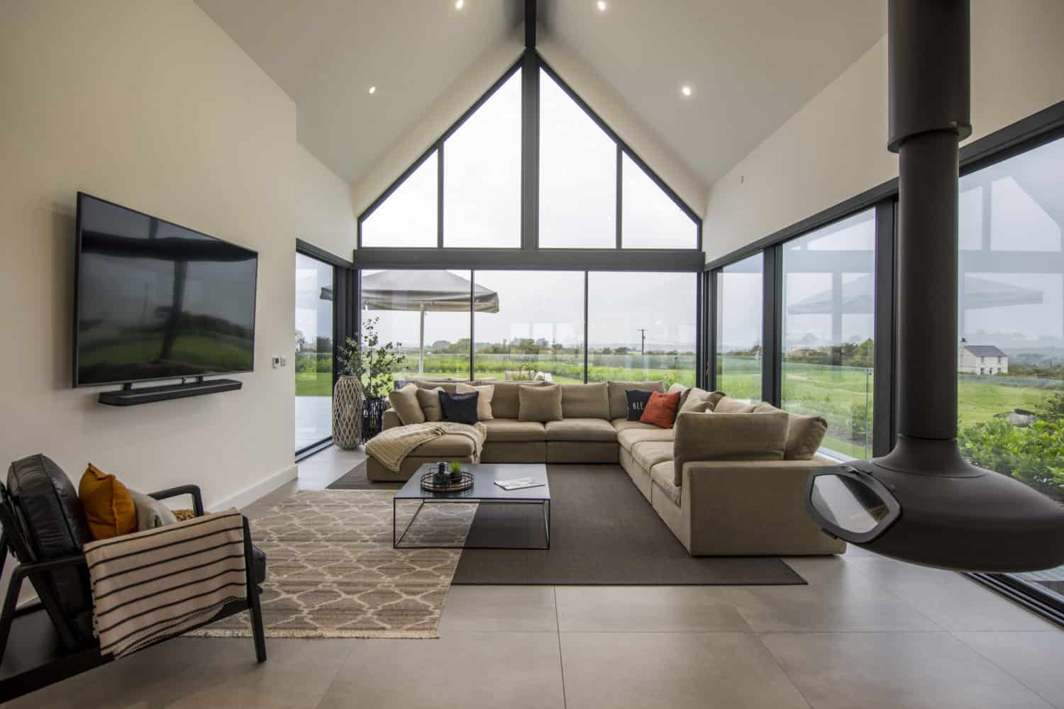 Modern living room with a beige sectional sofa, large windows offering scenic countryside views, a wall-mounted TV, contemporary fireplace, bifold doors, and minimalistic decor. Natural light fills the spacious, open area.
