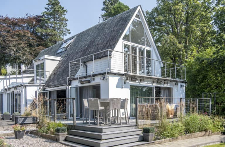 Modern two-story house with large glass windows and sliding doors, a sloped roof, and an upper-level balcony. Outdoor patio features a dining table and chairs, surrounded by plants and trees on a sunny day.