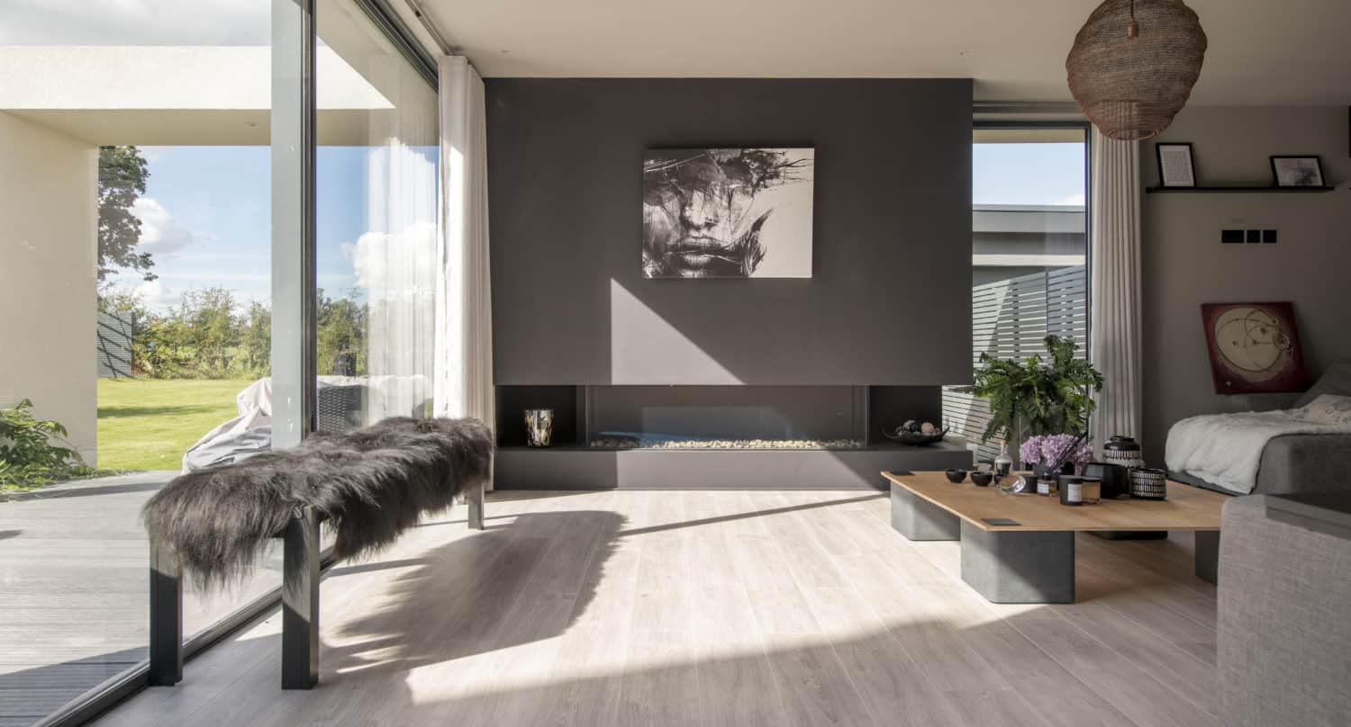 Modern living room with large windows, light wood floors, a dark accent wall with abstract art, a low wooden coffee table, a bench with fur cover, and sliding doors opening to views of the garden outside.