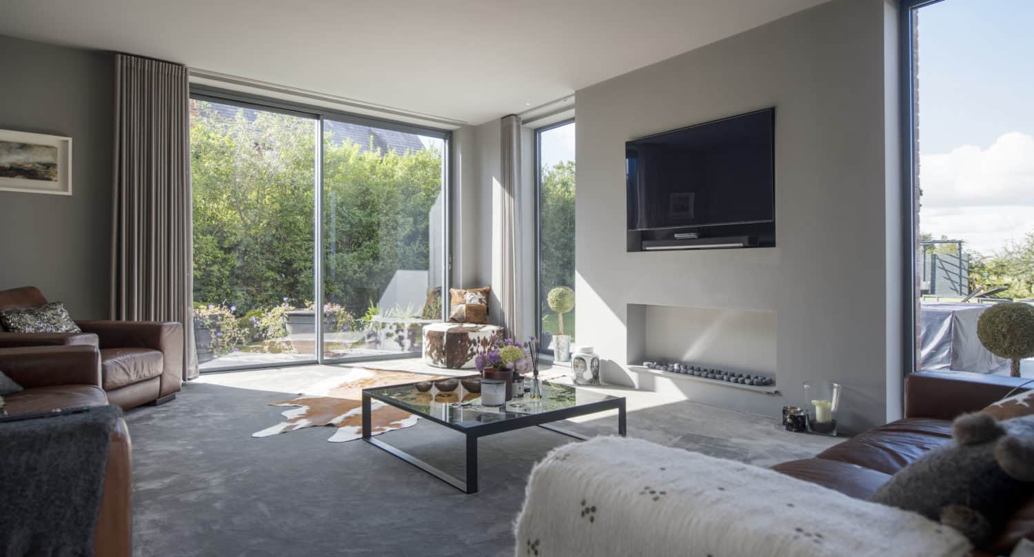 Modern living room with large windows, brown leather sofas, a coffee table, wall-mounted TV, and neutral decor. Sunlight fills the space, highlighting the greenery visible through bespoke glass solutions and sliding doors.
