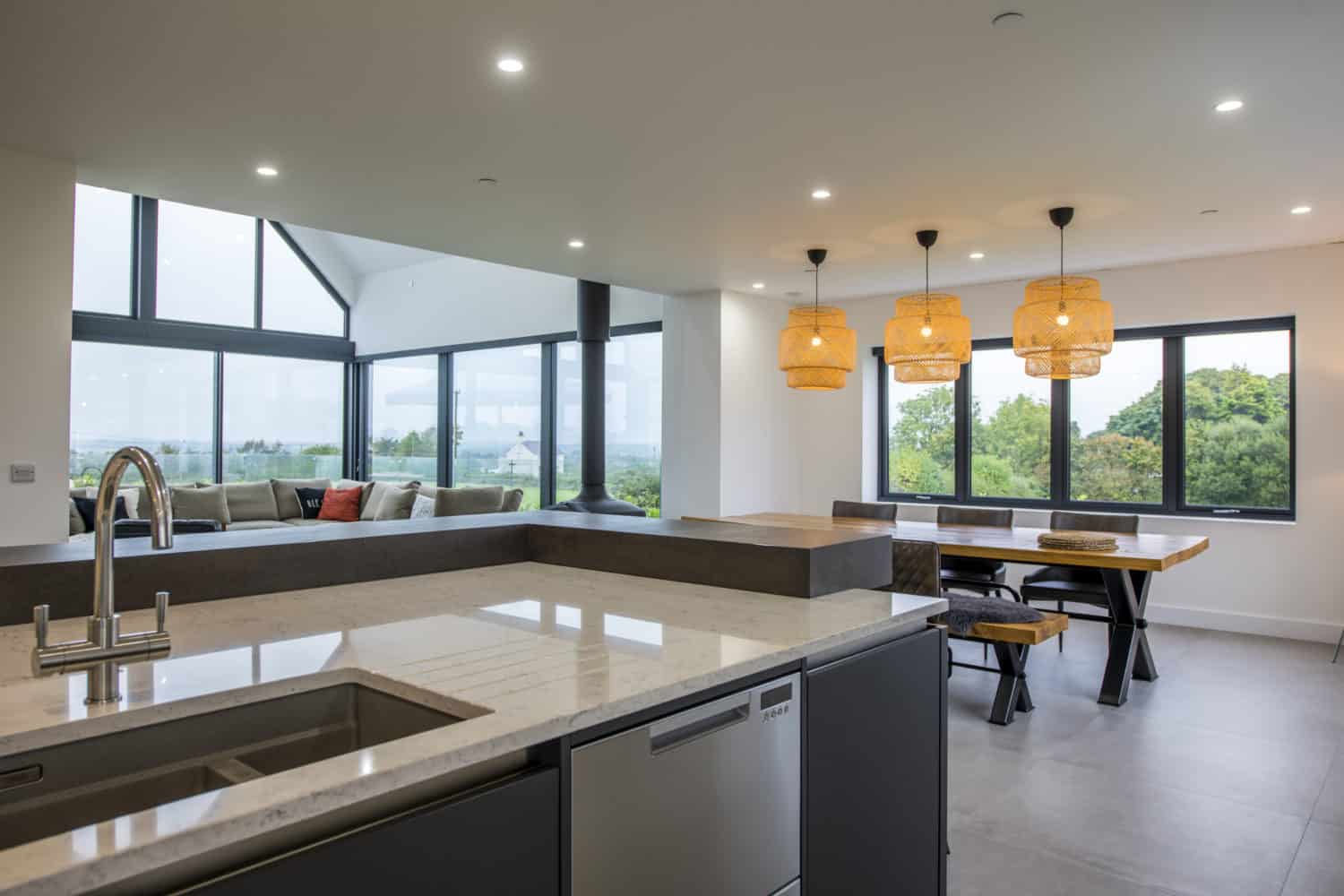 Modern open-plan kitchen and dining area with marble countertops, a sink, built-in dishwasher, wooden table, benches, and three woven pendant lights. Large bifold doors offer seamless views of greenery outside.