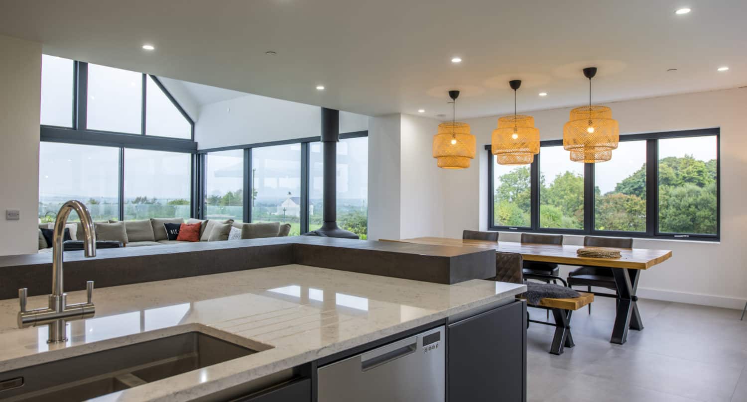 Modern open-plan kitchen and dining area with marble countertops, a sink, built-in dishwasher, wooden table, benches, and three woven pendant lights. Large bifold doors offer seamless views of greenery outside.