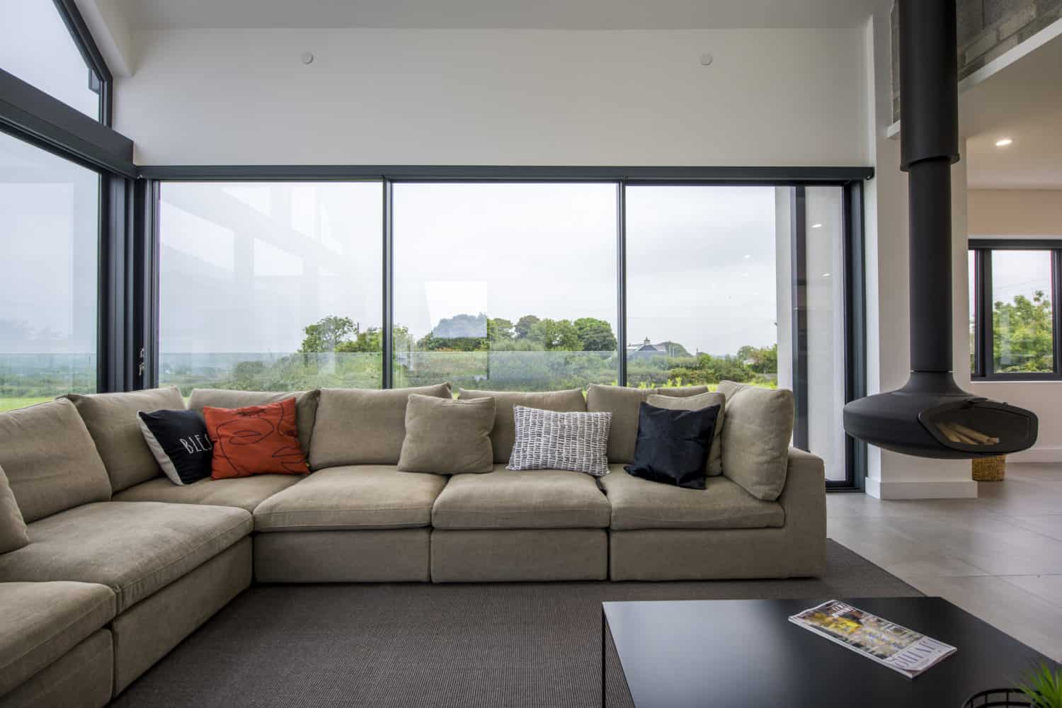 Modern living room with a large beige sectional sofa, decorative pillows, a black suspended fireplace, bifold doors, floor-to-ceiling windows, and views of green trees and countryside outside.
