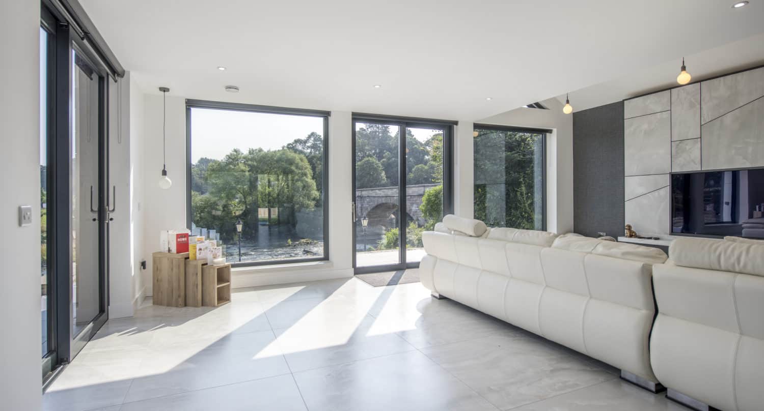 Modern living room with large floor-to-ceiling windows, white sectional sofa, marble tile floor, and natural light streaming in from sliding doors. Trees and a stone bridge are visible outside. Minimalist décor with a small wooden shelf.