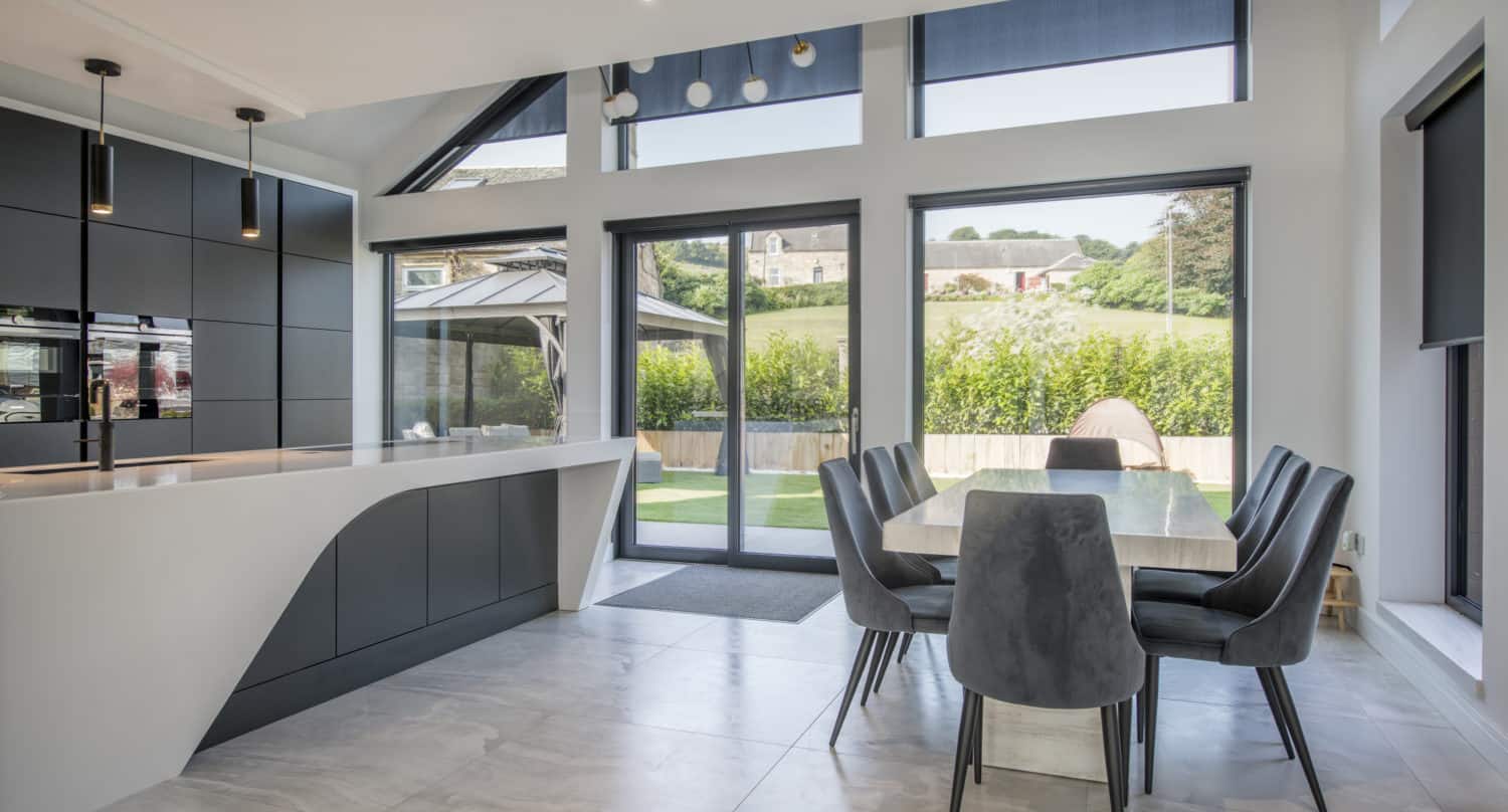 Modern kitchen and dining area with sleek black cabinets, a white island, grey chairs around a light wood table, large windows, sliding doors, and views of a green yard and countryside outside.