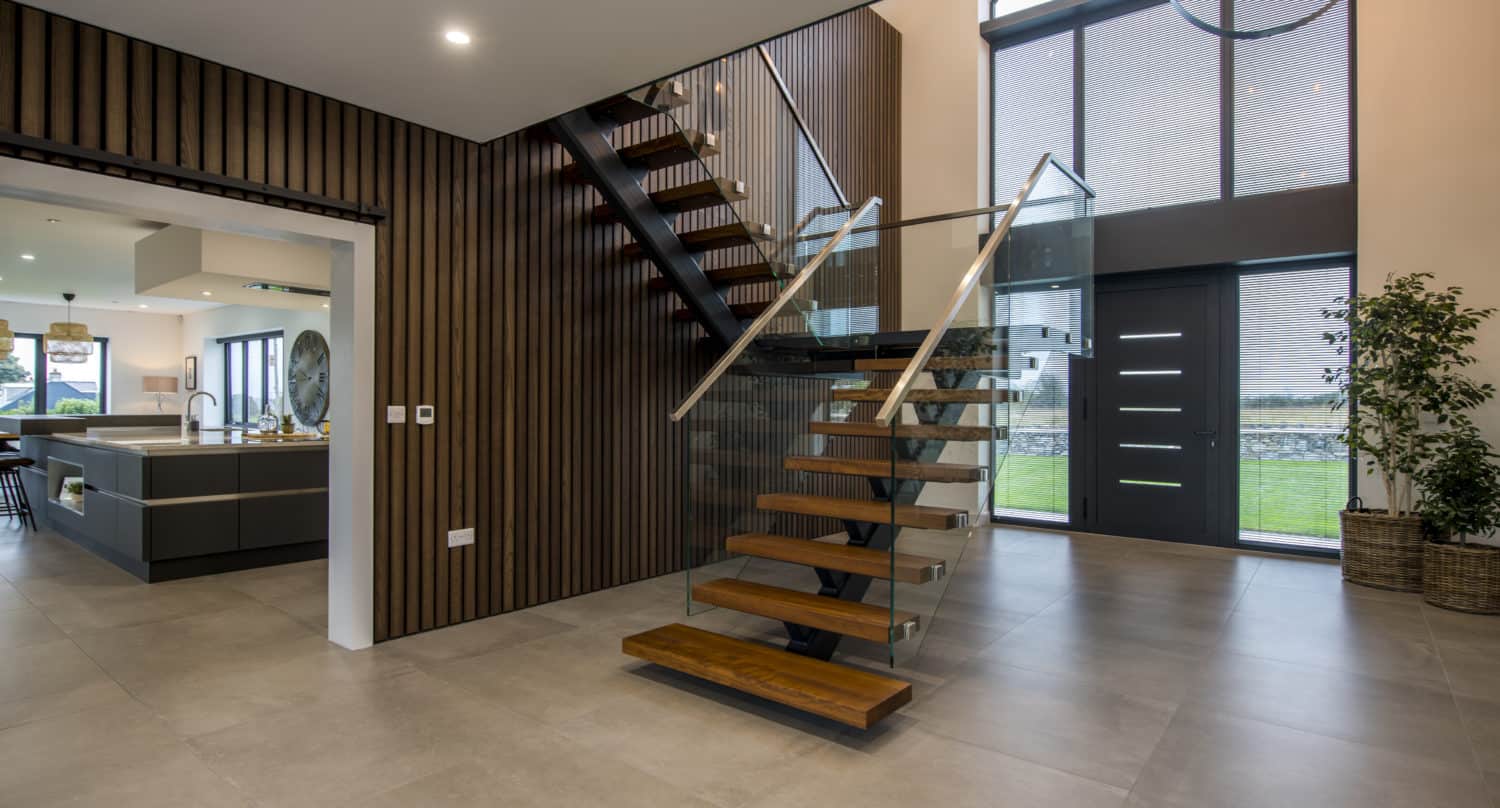 Modern open-plan foyer with floating wooden staircase, bespoke glass solutions in the railing, large windows, double front doors, tiled floor, wood-paneled wall, and view into a stylish kitchen. Indoor plants add a touch of greenery.