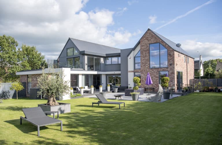 Modern two-story house with large windows, brick and white exterior, plus sleek sliding doors opening to a green lawn. Patio furniture, lounge chairs, potted plants, and a purple umbrella are arranged in the garden.