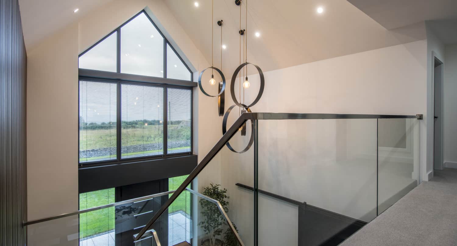 A modern home interior featuring bespoke glass solutions with a glass-railed staircase, large triangular windows, hanging circular pendant lights, and a view of a green lawn outside.