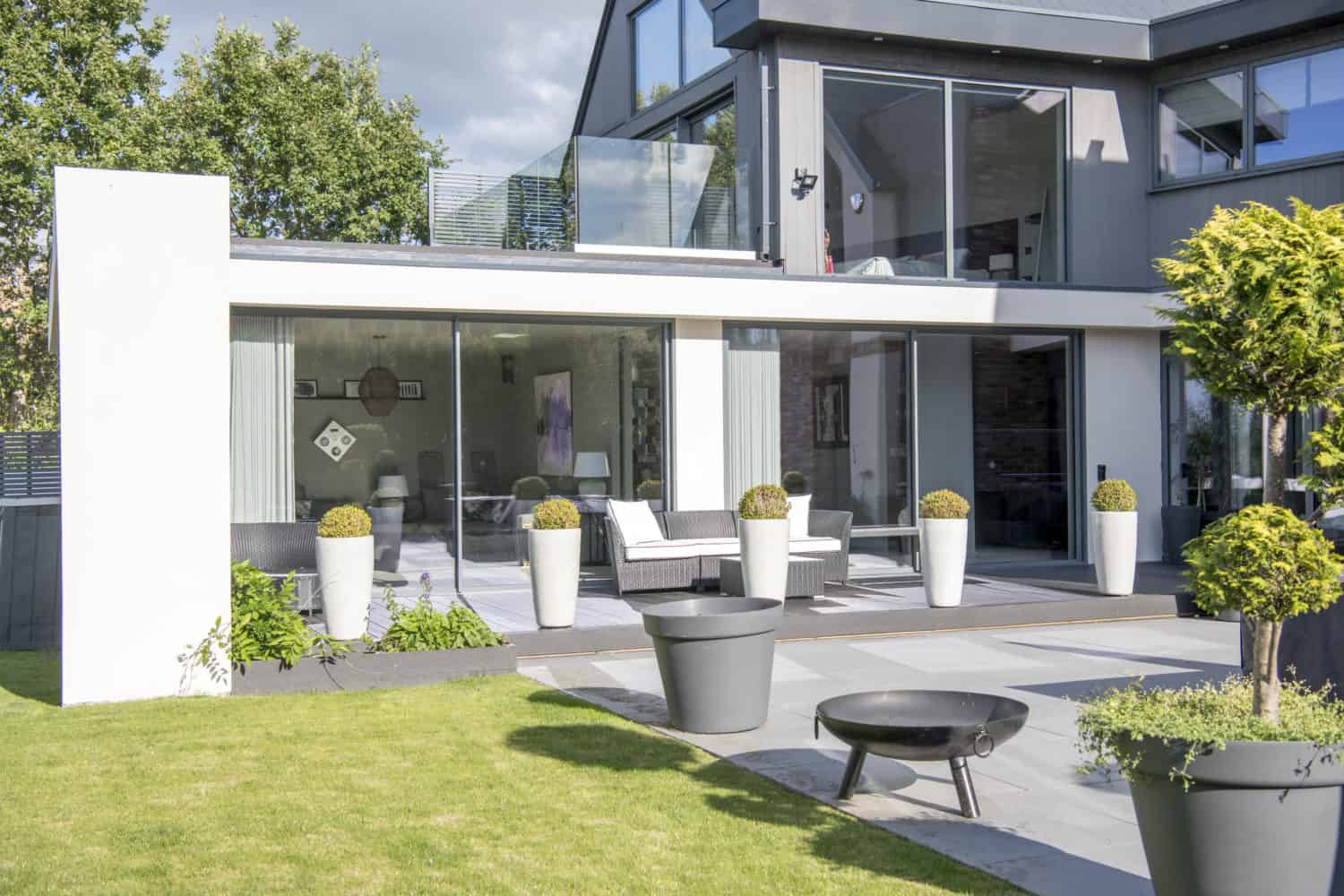 Modern house with large bifold doors, a patio with outdoor seating, tall white planters, a fire pit, and a neatly manicured lawn, surrounded by greenery and trees on a sunny day.