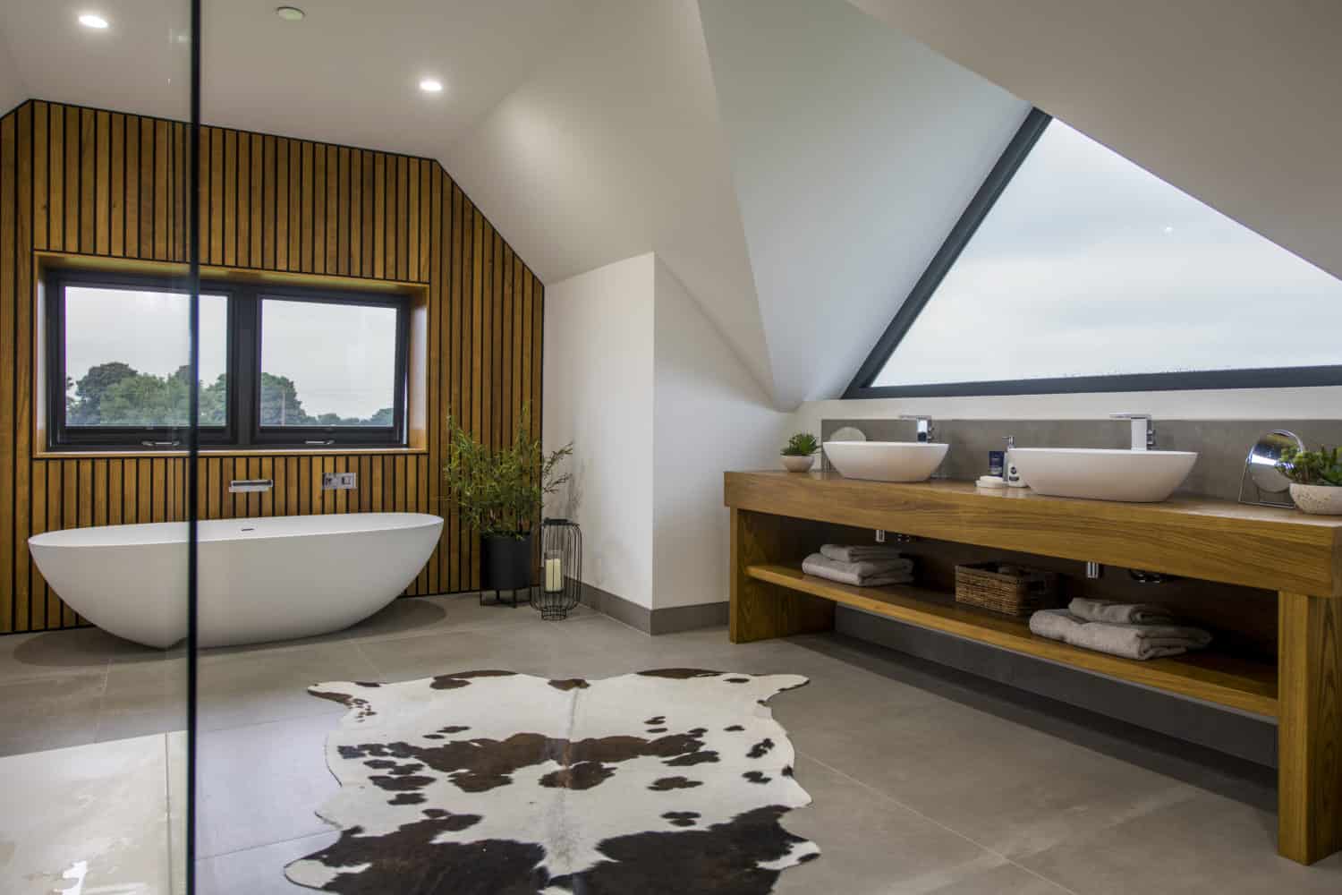 Modern bathroom with a freestanding white bathtub, double sinks on a wooden vanity, large triangular window, bifold doors, wooden slat wall, potted plants, and a cowhide rug on the floor.