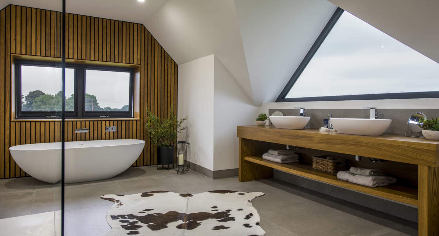 Modern bathroom with a freestanding white bathtub, double sinks on a wooden vanity, large triangular window, bifold doors, wooden slat wall, potted plants, and a cowhide rug on the floor.