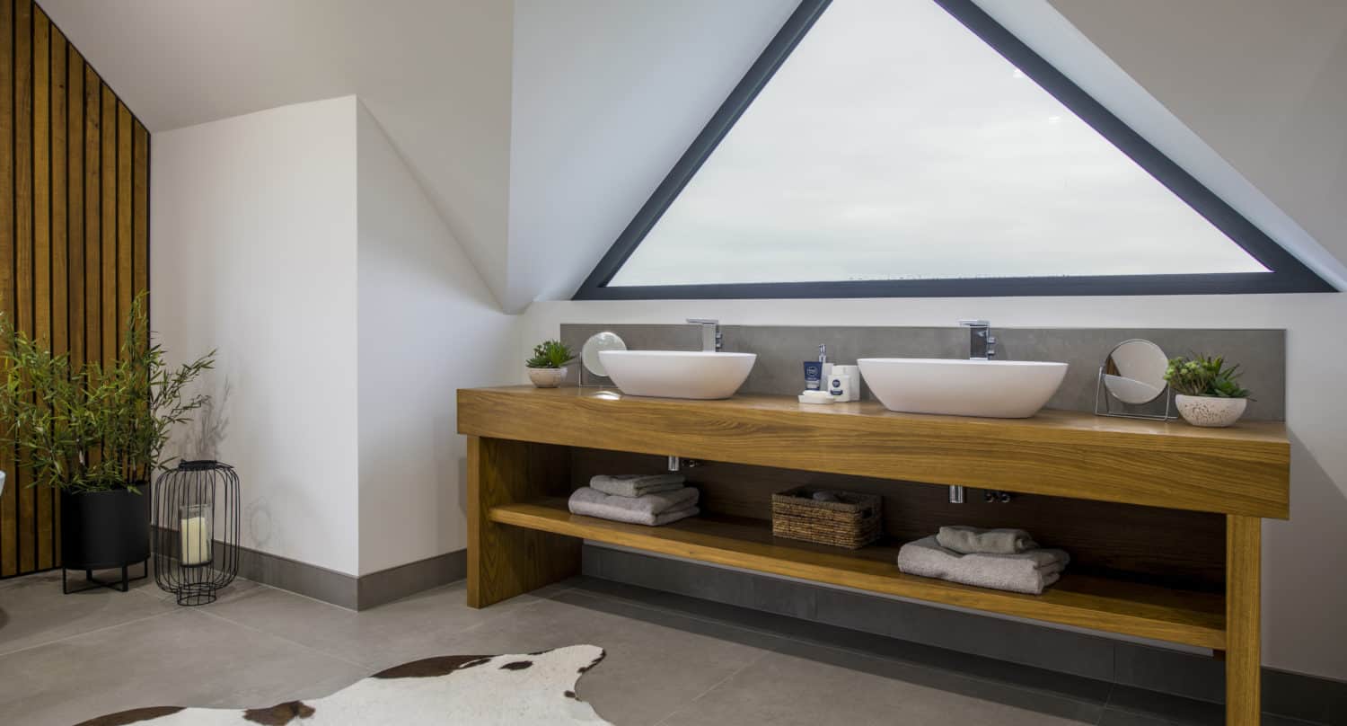 Modern bathroom with a wooden double vanity, vessel sinks, triangular window, potted plants, and neatly folded towels. Sliding doors offer privacy while a cowhide rug and bespoke glass solutions add style to the gray tile floor and tall corner plant.