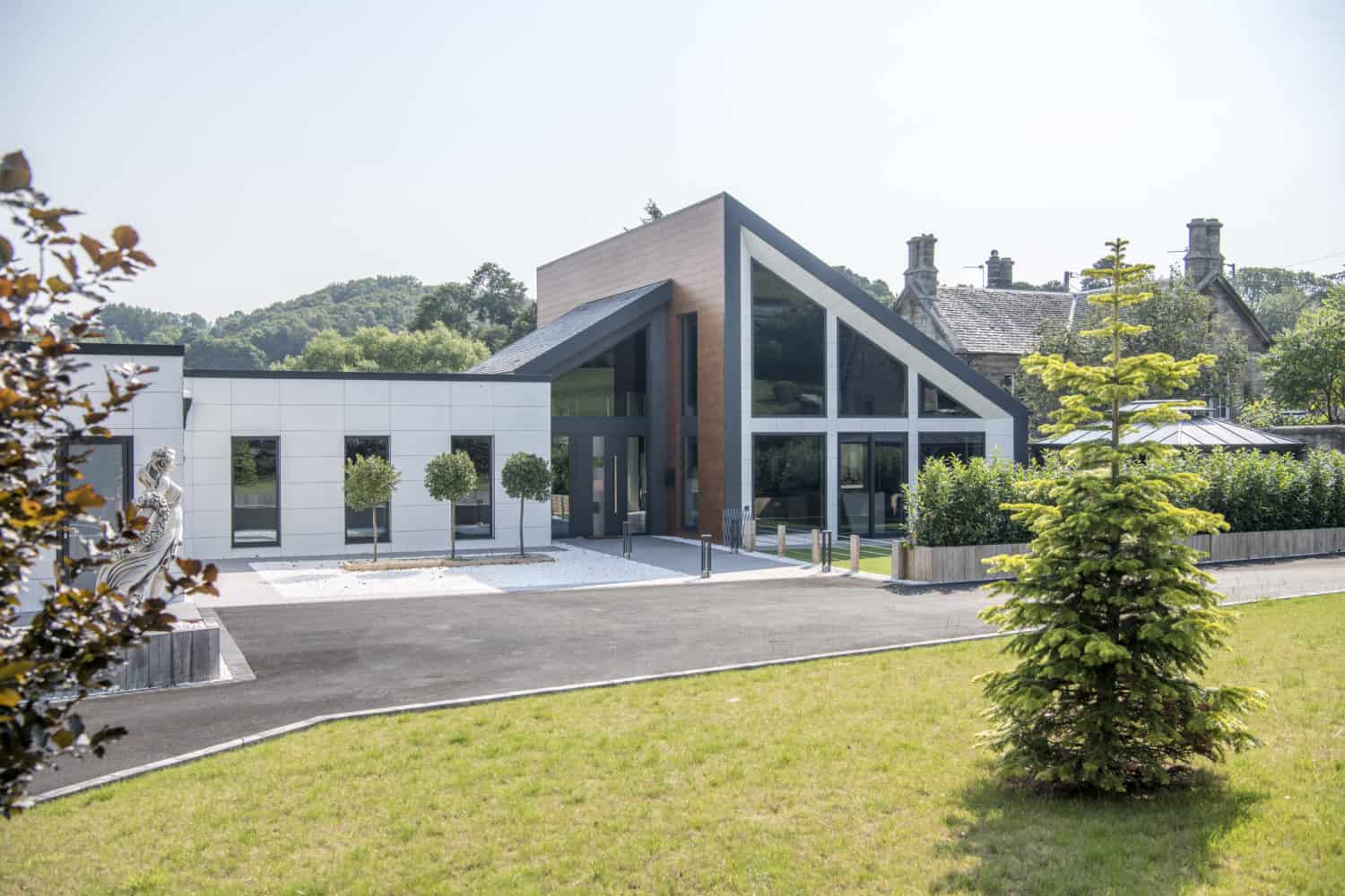 A modern building with large glass windows, sliding doors, and a slanted roof stands next to an older stone building, surrounded by green grass, trees, and landscaped shrubs on a sunny day.