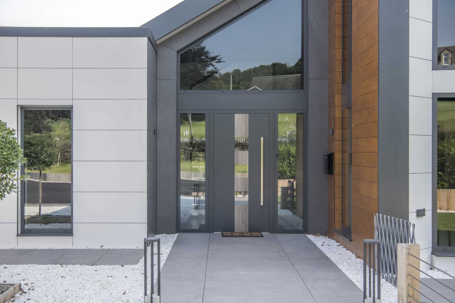 Modern house entrance with bespoke glass solutions, featuring large glass double front doors, vertical windows, and a mix of white, gray, and wood paneling. The pathway is bordered by white gravel and a metal fence. Green hills are visible in the reflection.