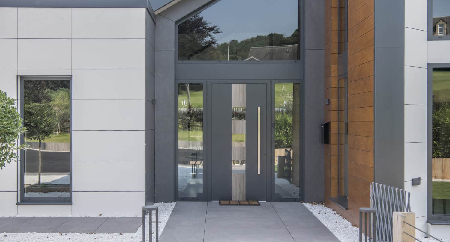 Modern house entrance with bespoke glass solutions, featuring large glass double front doors, vertical windows, and a mix of white, gray, and wood paneling. The pathway is bordered by white gravel and a metal fence. Green hills are visible in the reflection.