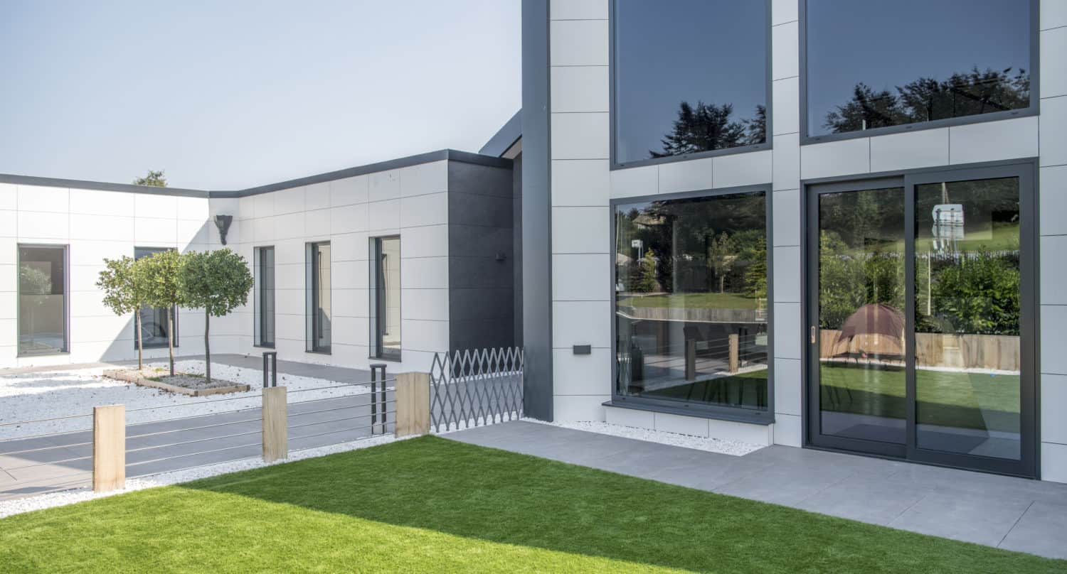 Modern house exterior with large glass windows, white walls, bifold doors, a small tree, neatly trimmed grass lawn, and a minimalist fence in a bright, sunny setting.