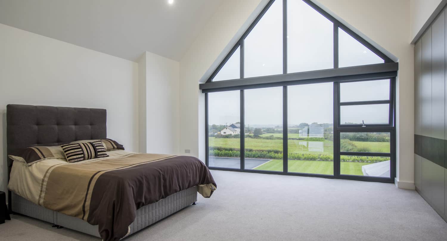 Modern bedroom with a double bed, brown and beige bedding, and large triangular floor-to-ceiling bifold doors offering a scenic view of green fields and distant houses. The room has white walls and a light carpet.
