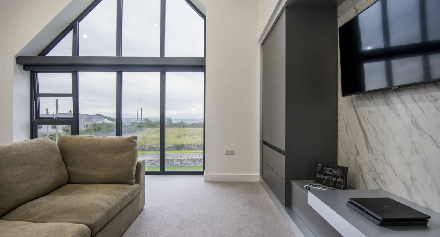 Modern living room with a beige sofa, large floor-to-ceiling windows and glass roofing offering a scenic outdoor view, a wall-mounted TV, marble accent wall, and a built-in shelf with electronic devices.