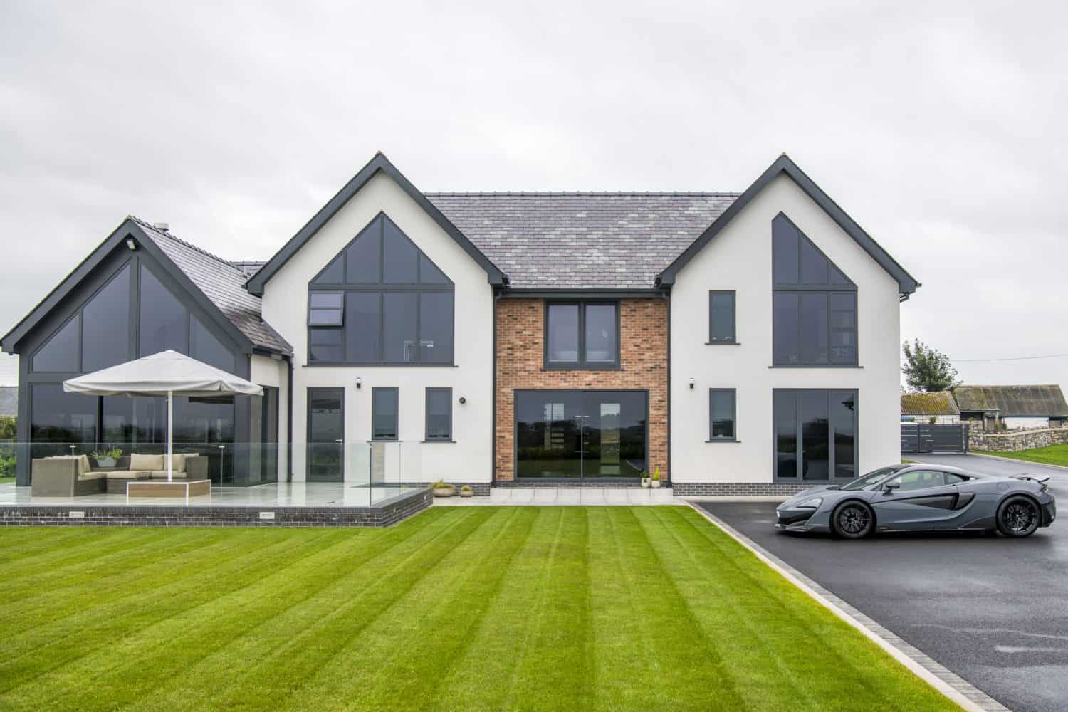 Modern two-story house with large windows and a mix of white and brick exterior. Stylish bifold doors open onto a patio with outdoor furniture, while a sports car is parked in the driveway and the neatly mowed lawn completes the look.