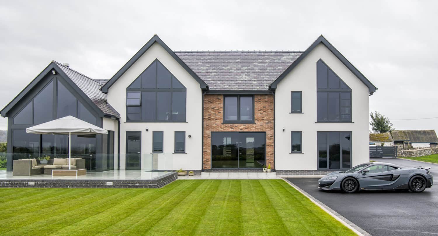 Modern two-story house with large windows and a mix of white and brick exterior. Stylish bifold doors open onto a patio with outdoor furniture, while a sports car is parked in the driveway and the neatly mowed lawn completes the look.