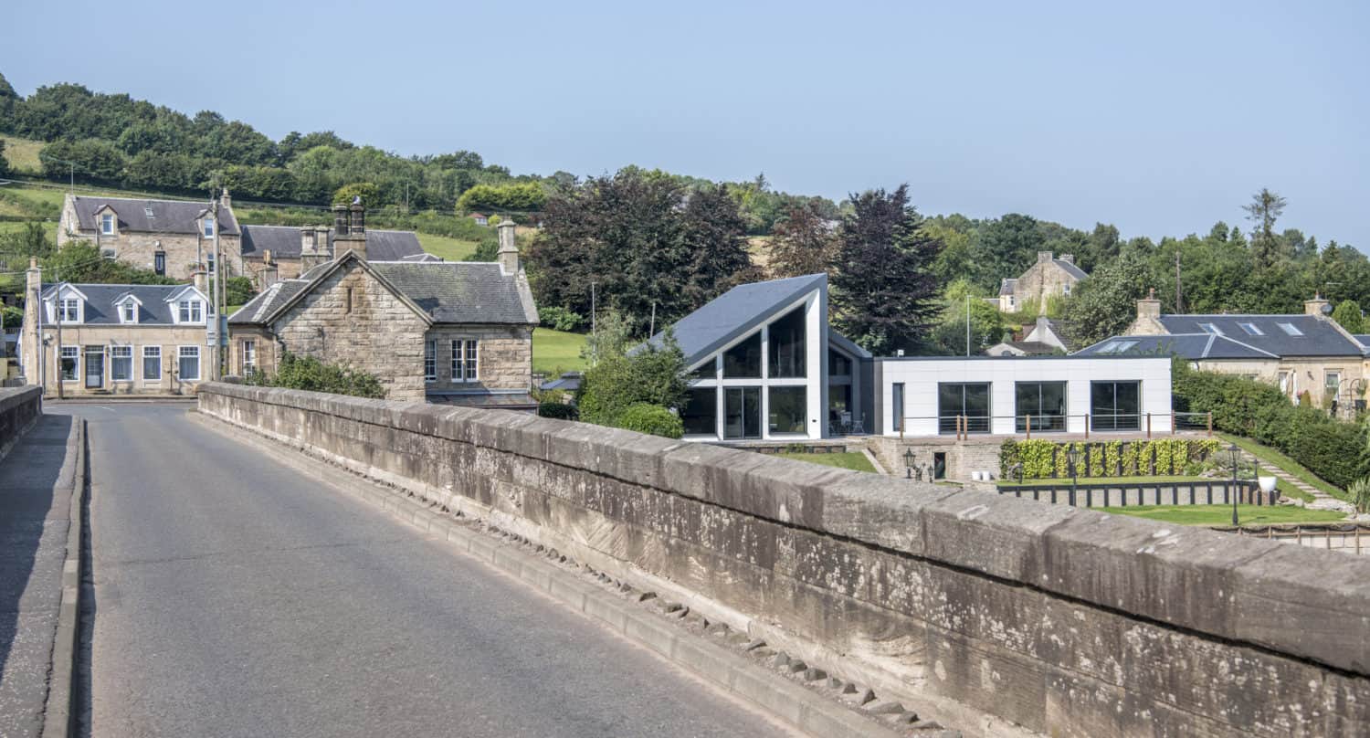 A stone bridge leads to a village with traditional stone buildings and a modern house featuring sleek sliding doors, set among green hills and trees under a clear blue sky.