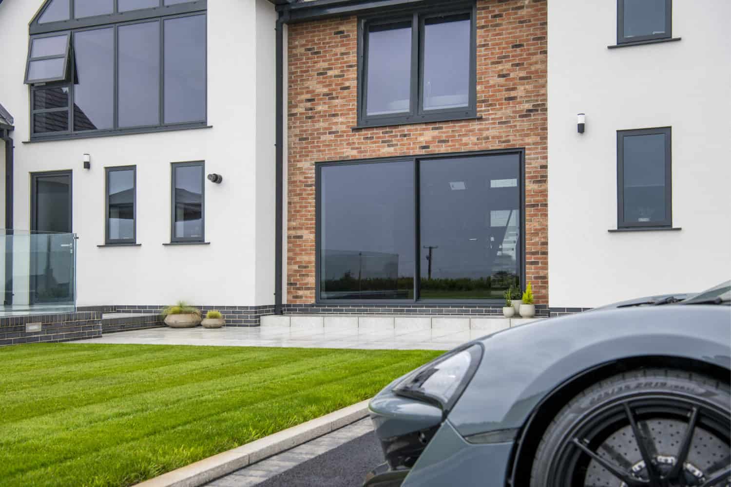 A modern house with large windows, bespoke glass solutions, a white and red brick exterior, neatly trimmed green lawn, and part of a sleek gray sports car parked in the driveway in the foreground.