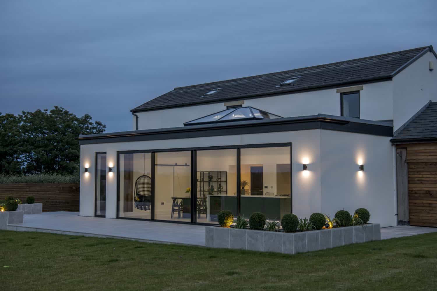 Modern two-story house with large glass windows and sleek bifold doors, illuminated outdoor wall lights, neatly trimmed bushes, patio, and a spacious lawn in the foreground at dusk.