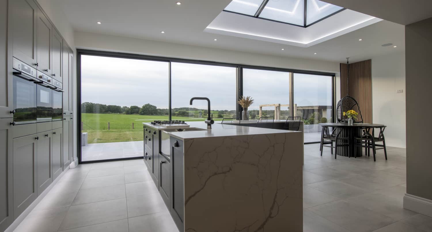 Modern kitchen with marble island, built-in appliances, large skylight, and bespoke glass solutions like floor-to-ceiling sliding doors opening to a scenic green landscape. Dining area with table and chairs is visible in the background.