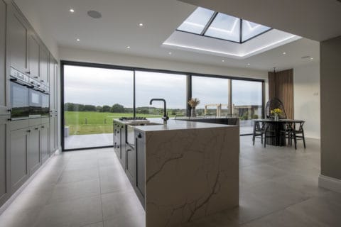 Modern kitchen with marble island, built-in appliances, large skylight, and bespoke glass solutions like floor-to-ceiling sliding doors opening to a scenic green landscape. Dining area with table and chairs is visible in the background.