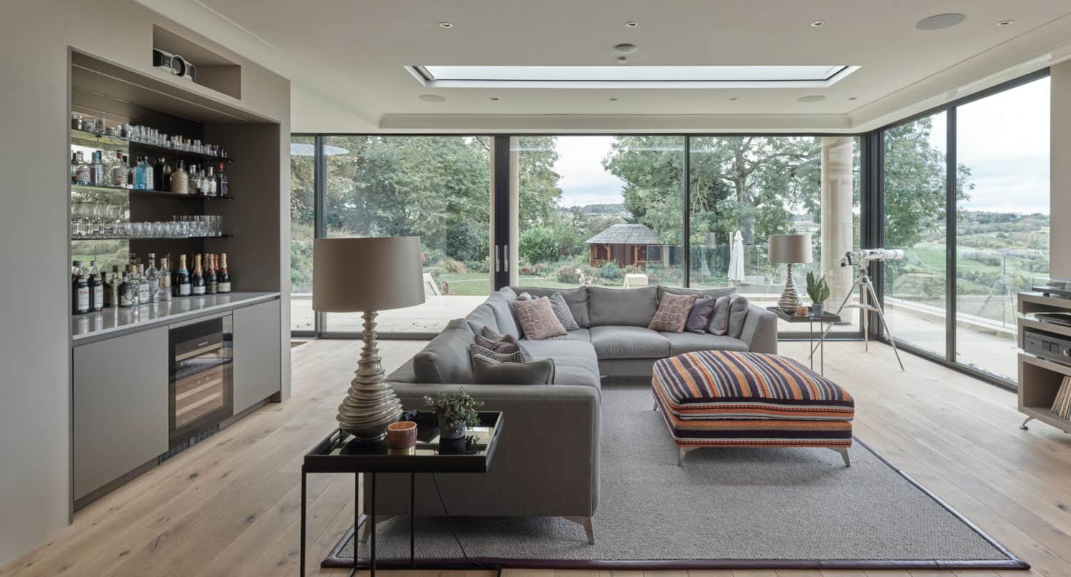 Modern living room with large glass windows, sliding doors, a gray sectional sofa with colorful pillows, a striped ottoman, built-in bar area, side tables with lamps, wooden floor, and views of trees and countryside outside.