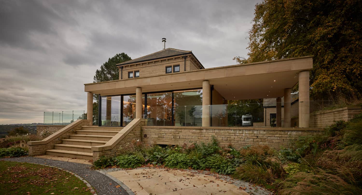 A modern house with large glass windows and stone walls sits on a landscaped hill, featuring sliding doors to a covered patio area, wide steps, and surrounding greenery under a cloudy sky.