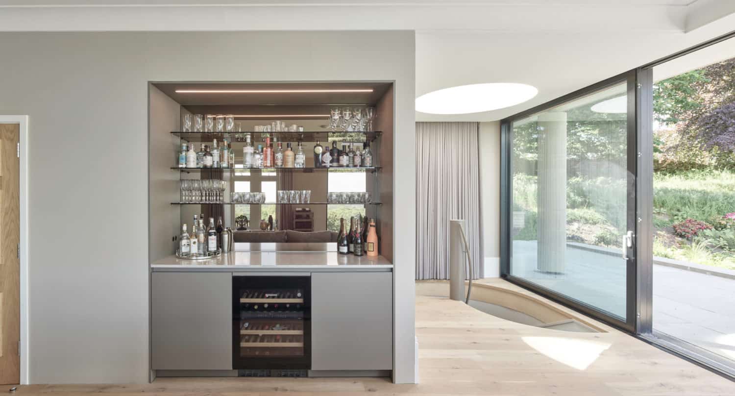 A modern home bar with shelves of glasses and bottles, a countertop with more bottles, and a built-in wine fridge below. The airy room features large windows and bespoke glass solutions overlooking the patio and garden.