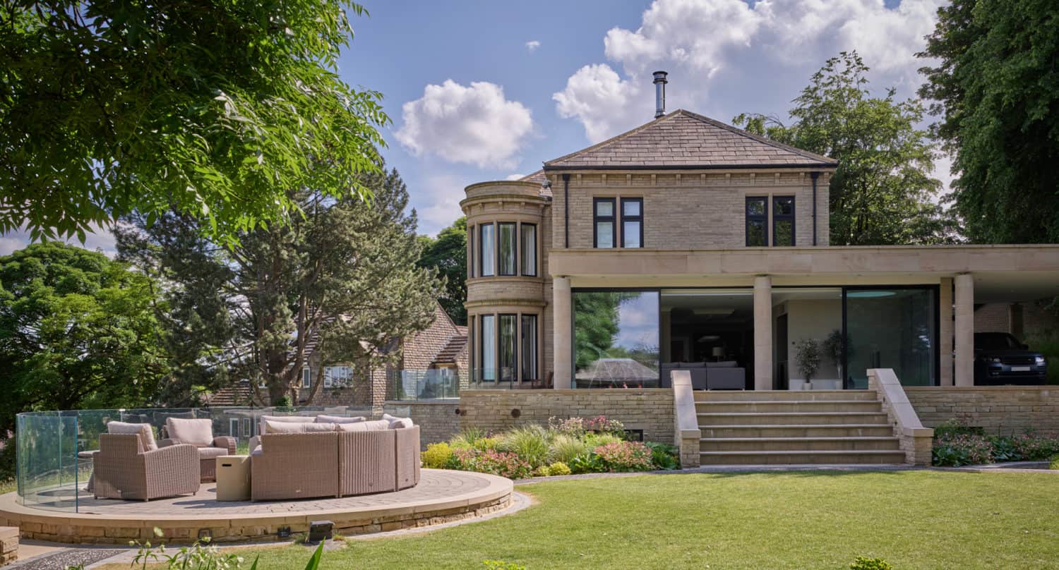 A modern two-story house with large windows, bespoke glass solutions, a stone exterior, and a circular outdoor seating area on a lawn, surrounded by trees and landscaped gardens under a partly cloudy sky.
